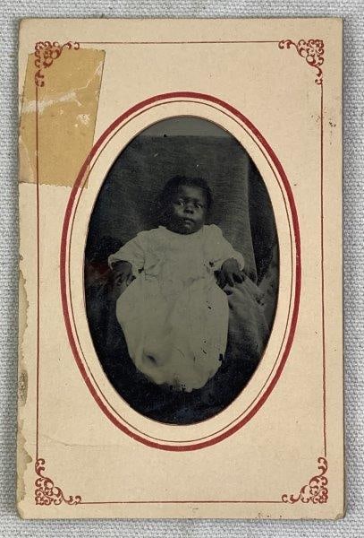 19th Century Photograph of African American Child: Touching portrait photograph depicting an African American child dressed in white, presented in a decorative sleeve with an oval cutout framing the image. Displays a warm aged patina. Measures approxi