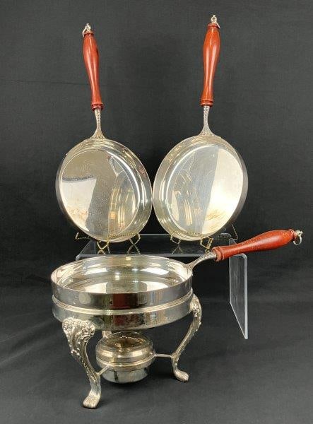 Three Italian Silverplate Crêpe Saucepans: A set of three Italian silverplate crêpe saucepans, each with a wooden handle and accompanied by an elegant footed warmer with ornate detailing. The pieces display rich silverplate tones and refi