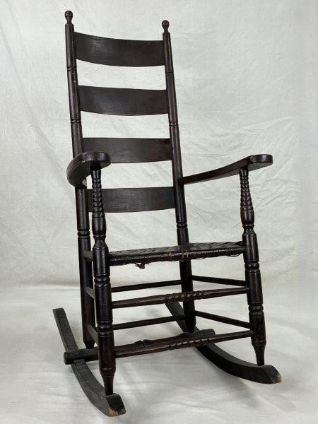 Painted Ladderback Rocking Chair: A traditional ladderback rocking chair with turned posts, woven seat, and a brown painted finish, exhibiting classic country form and sturdy construction. Measures approximately 48" H x 23 1/8" W x 31