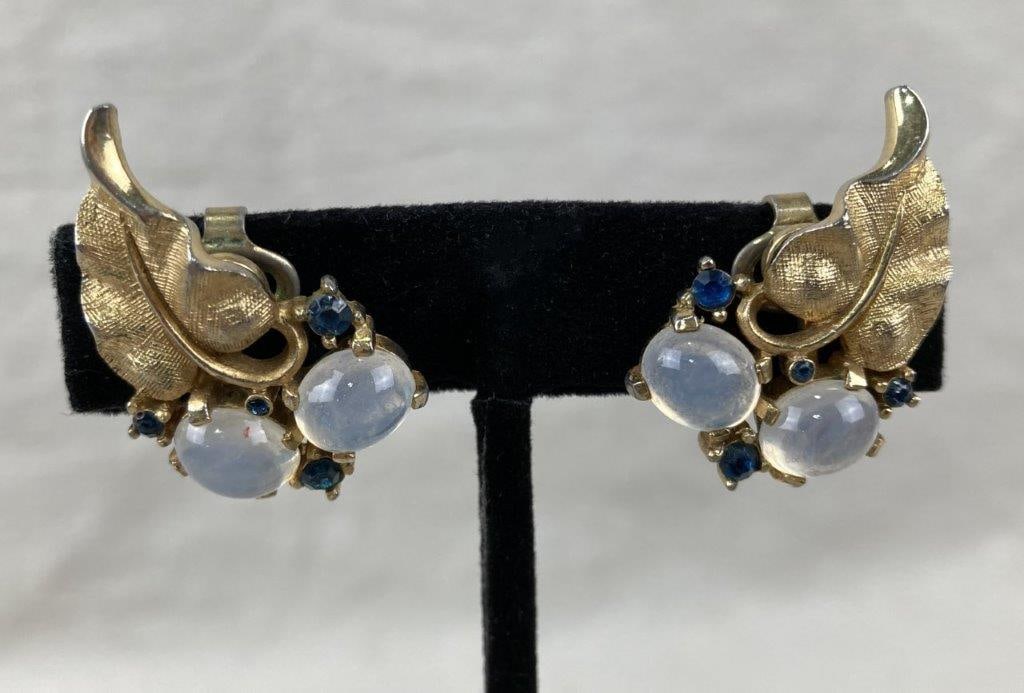 Vintage Trifari Gold-tone Leaf Earrings: Pair of vintage Trifari clip-on earrings featuring large clear stones accented with smaller blue stones and goldtone leaf motifs, offering classic mid-century glamour. Each earring bears the Trifari m