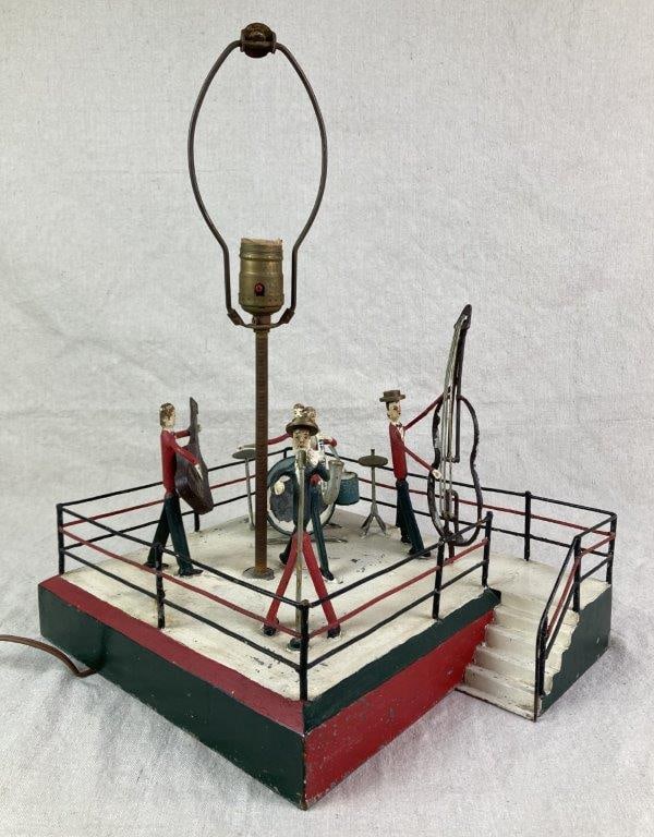 Folk Art Metal Musician Lamp: A folk art metal table lamp featuring a lively band scene arranged on a stage-form base with surrounding railing. Painted metal figures depict a singer in a green shirt alongside band members in red s