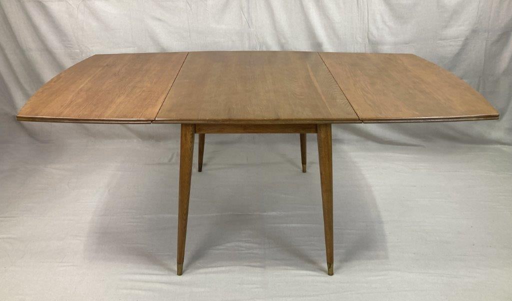 Mid-Century Modern Drop-Leaf Dining Table: A mid-century modern dining table featuring a rectangular top with rounded ends and tapered legs fitted with brass caps. Exhibits a warm, mellowed patina. With both leaves raised, the table measures