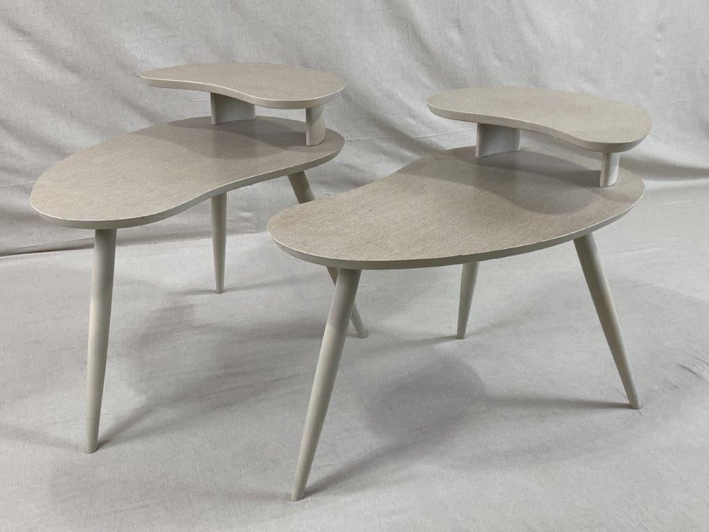 Pair of Mid Century Modern Sculpted table: A pair of Mid Century Modern two-tiered side tables featuring softly curving lines, light vanilla-toned finishes, and tapered legs characteristic of the period. The open lower tier adds both function