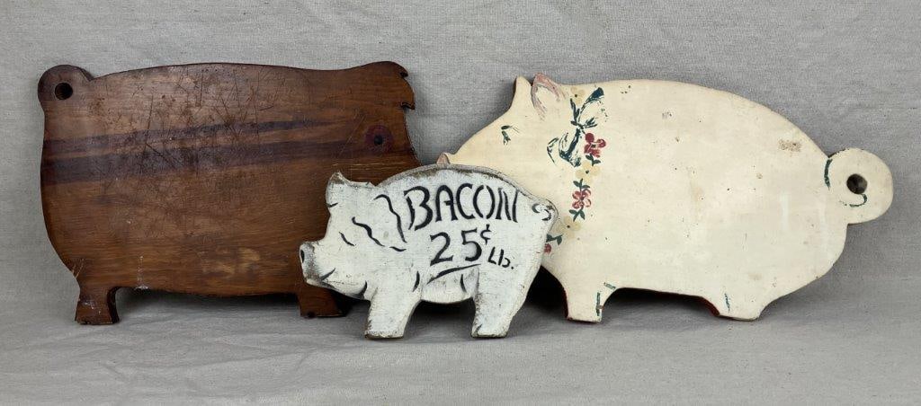 Three Wood Pig Cutting Boards: A group of three vintage pig-shaped wooden plaques, including a wall hanging with a white ground, red rim, and painted floral decoration; a white decorative pig-form sign reading “Bacon 25 cents