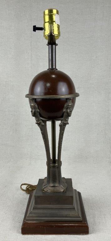 Robert Abbey Art Deco Style Lamp: An Art Deco–style table lamp by Robert Abbey, featuring a central spherical form supported by a brass tripod base adorned with ornamental face motifs and finished in a bronze tone. Measures