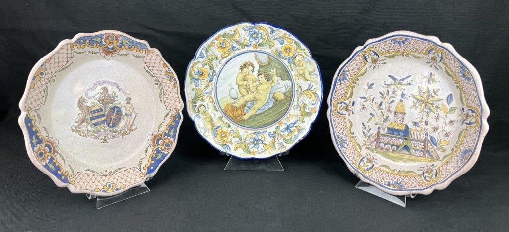 Three Majolica Plates: A group of three tin-glazed earthenware Majolica plates decorated with classic motifs. One plate depicts classical youths engaged in a Bacchanalian wine scene, another features a family crest, and