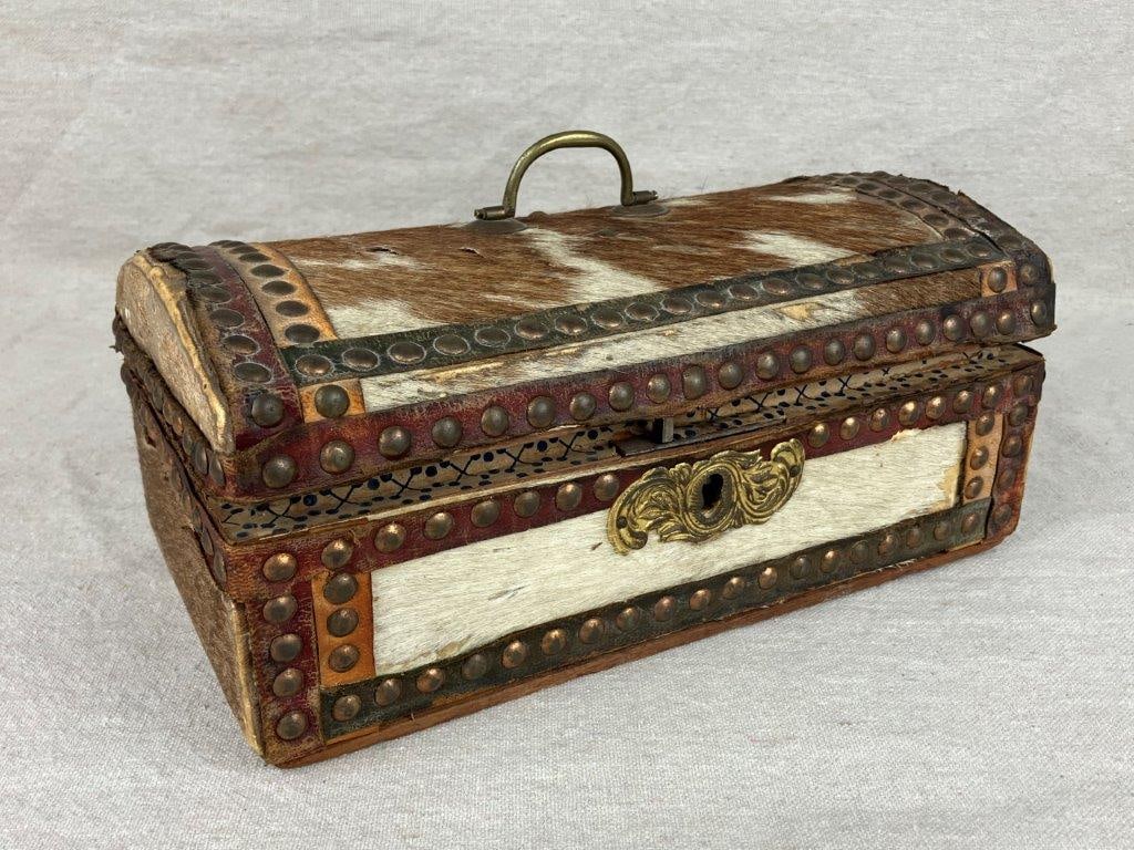 Cowhide-Covered Box: A rustic antique box covered in cowhide, fitted with brass hardware, brass studs, and a locking clasp. Measures approximately 5 1/4â€³ H x 10â€³ W x 5 3/4â€&
