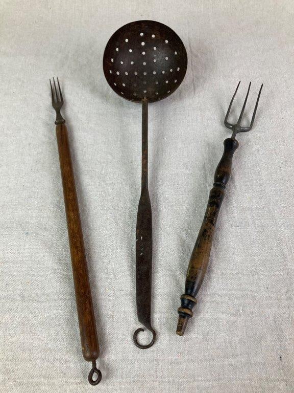 Kitchen Utensils: A small assortment of antique kitchen utensils, including a toasting fork with a turned wooden handle, a metal fork with a wooden handle, and a wrought iron strainer ladle. Exhibits a worn, aged
