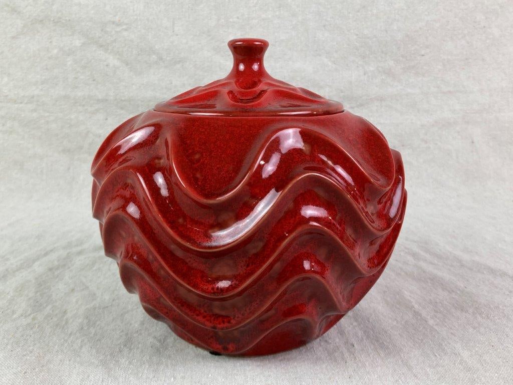Mid-Century Modern Ceramic Jar: A mid-century modern ceramic jar finished in a deep red glaze, decorated with a rhythmic wave pattern. Designed with a knobbed lid and fitted with a metal insert. Measures approximately 7 1/2″