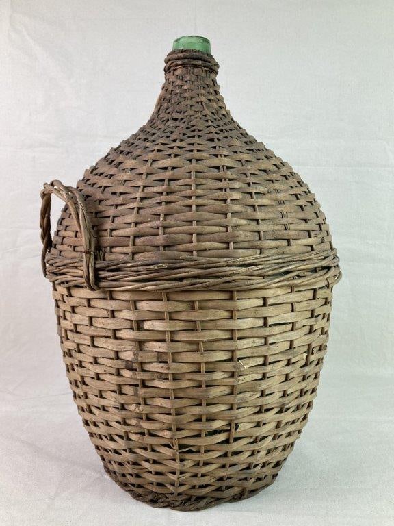 Wicker Wrapped Demijohn: An antique demijohn with a large green hand-blown glass body encased in protective woven wicker, featuring integrated carrying handles. Displays a worn, aged patina consistent with use. Measures appro