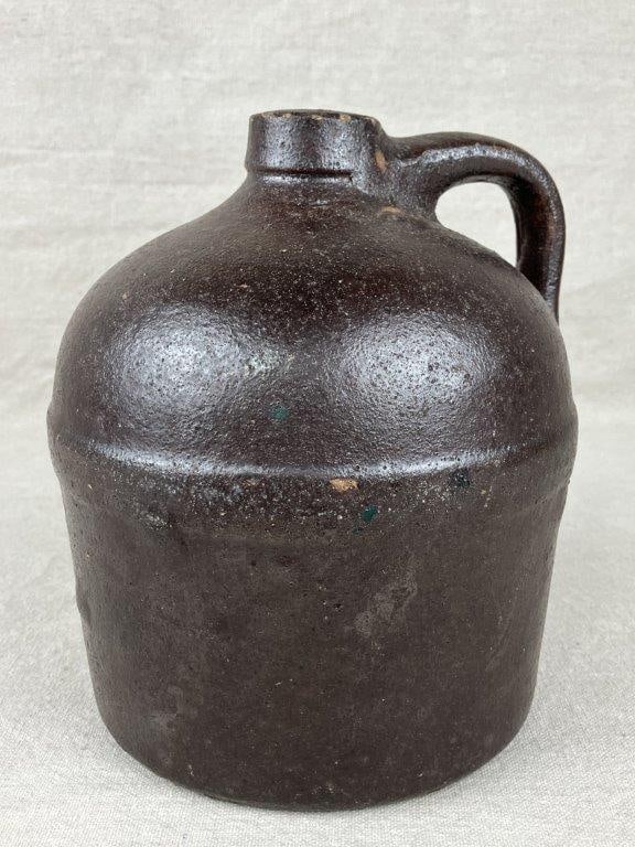 Glazed Stoneware Jug: A glazed stoneware jug with an applied handle, finished in a rich, deep brown glaze. Measures approximately 8″ H x 6 1/2″ in diameter. Shows age-appropriate wear consistent with use. Some
