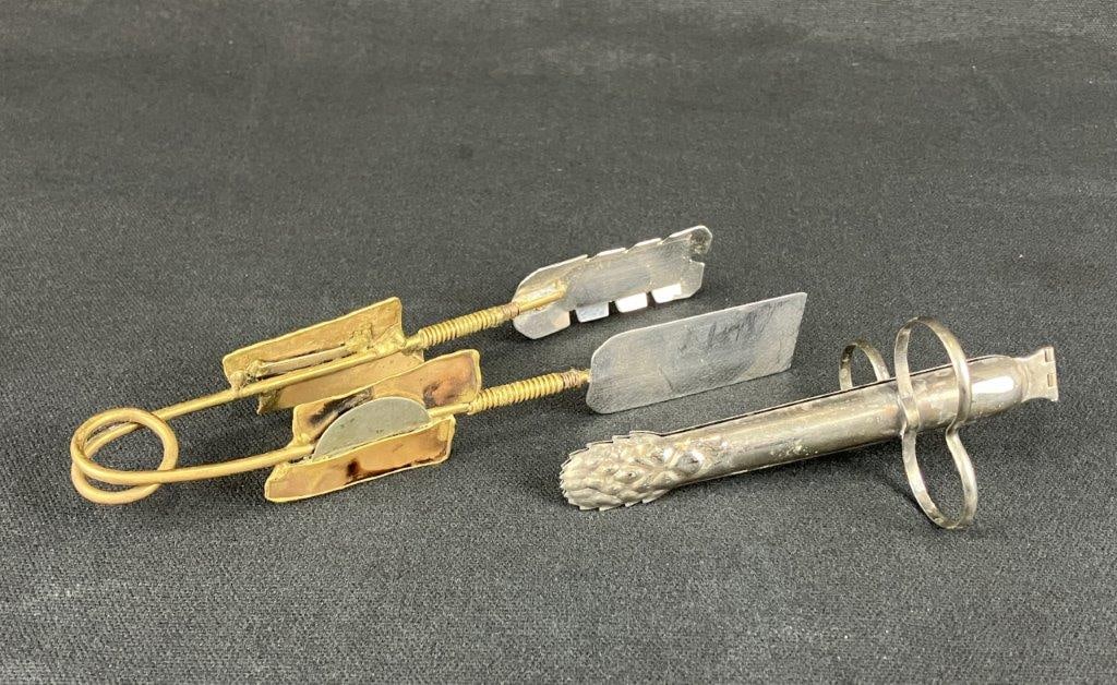 Two Sugar Tongs Including Mixed Metals: A pair of sugar tongs in mixed metals, including one artisan-crafted example with a hand-formed design. The handcrafted tongs measure approximately 1 1/4″ H x 7 1/8″ W x 1″ D. In