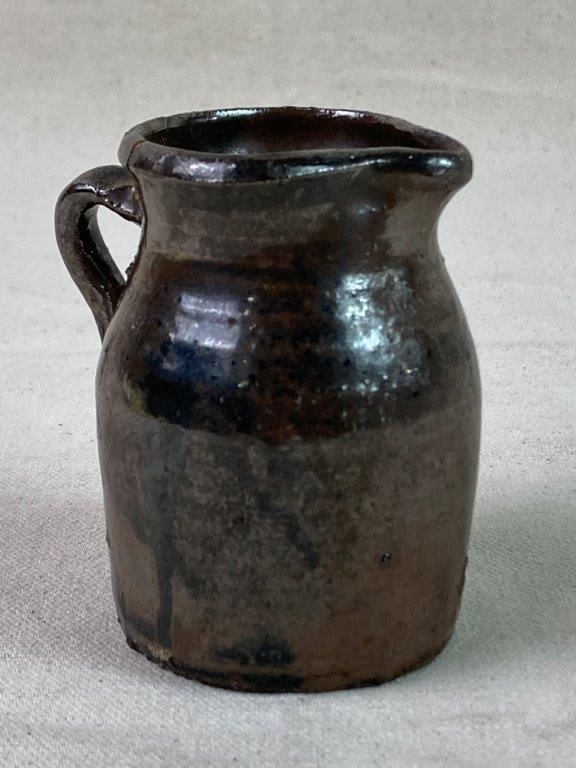 Small Stoneware Pitcher: A small stoneware pitcher with a brown glaze and applied handle, exhibiting a worn, aged patina. Measures approximately 3â€³ H x 2â€³ W x 2 1/2â€³ D. Sh