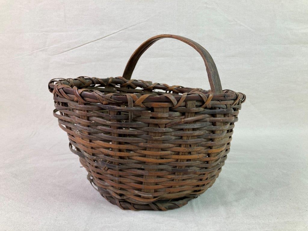 Country Gathering Basket: A nicely woven rivercane gathering basket with a wrapped rim, arched handle, and round wrapped base. Displays a worn, aged patina from use. Measures approximately 8 5/8″ H x 8 3/4″ W x