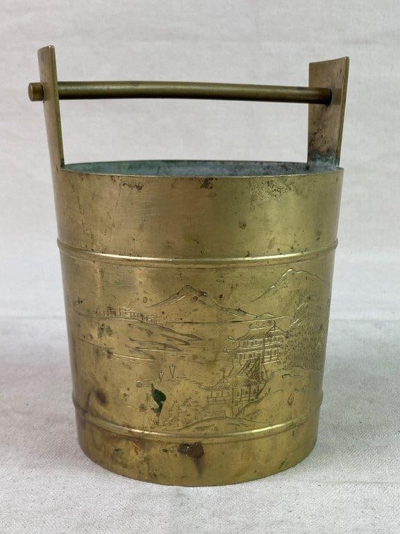 Vintage Solid Brass Bucket: A vintage solid brass bucket with a swing handle, etched with a Chinese landscape scene depicting mountains and architectural elements. Exhibits a worn, aged patina. Measures approximately 6