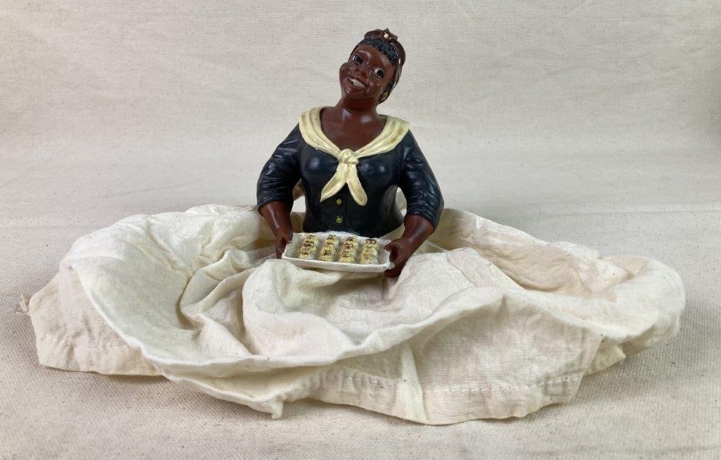 Black Americana Porcelain Doll: A porcelain doll depicting an African American woman holding baked goods on a tray. She wears a red-and-white polka dot bandana and a white fabric skirt. Exhibits a worn, aged patina. Measures