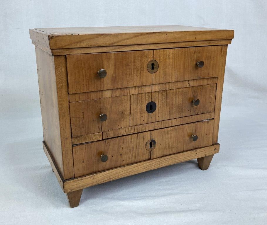 Miniature Biedermeier Fruitwood Chest: 19th Century, a finely constructed miniature chest featuring three long drawers accented with elegant brass knobs and inset escutcheon keyholes. Crafted from fruitwood, the piece is supported by