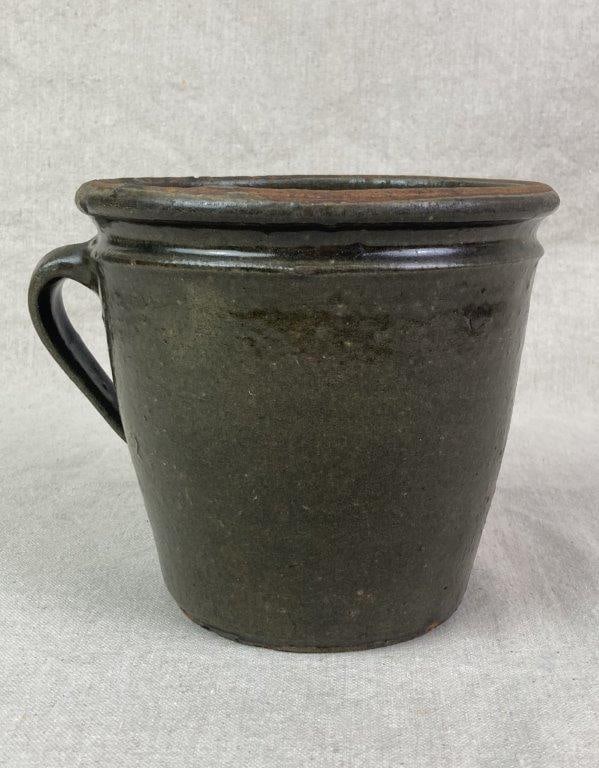 Edgefield Clabber Pot: A stoneware clabber pot with a dark olive-brown alkaline glaze, featuring a tapering cylindrical body with a heavily rolled rim and applied handle, retaining a worn patina consistent with age and