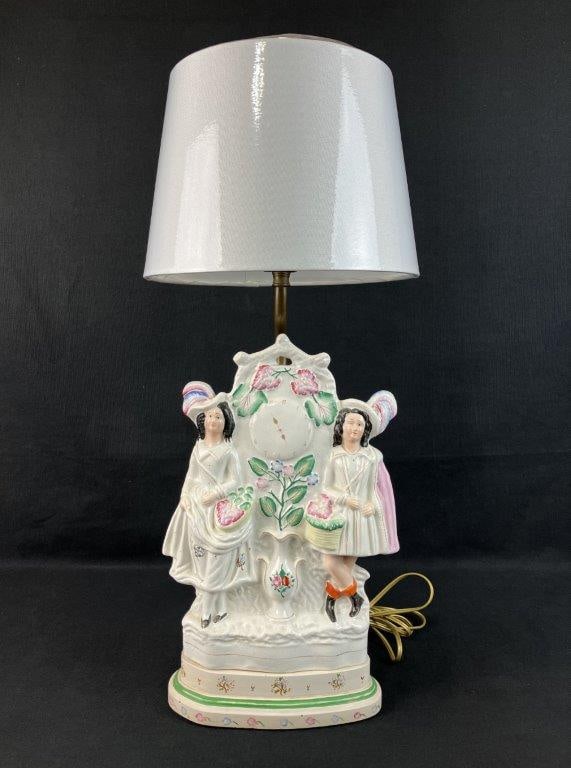 Staffordshire Courting Couple Figurine Lamp: Depicts a couple in classic English dress flanking a central clock form adorned with floral details, likely representing a courting man and woman. Creamy white ground with pink and green accents and
