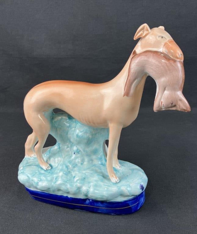 Staffordshire Whippet: Elegant Staffordshire figure depicting a whippet holding a rabbit as a hunter's bounty. Decorated in soft aqua, blue, and orange-brown tones throughout. Measures approximately 10" H x 8 1/2" W x 2