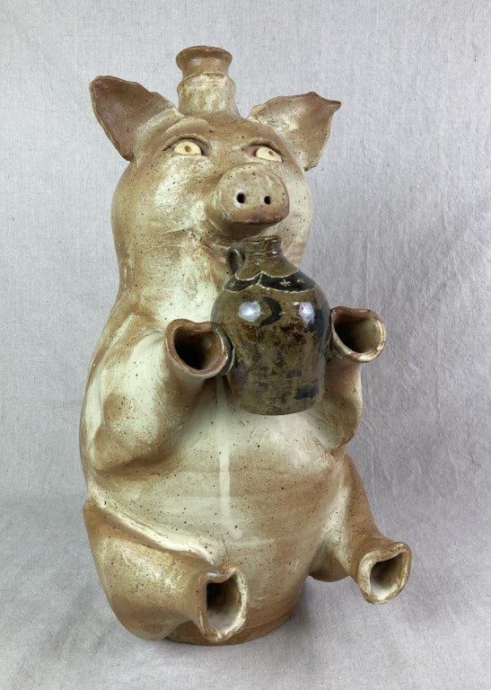 Edgefield Pig Jug by Bob Taft: Large speckled glaze stoneware jug sculpted in the form of a seated pig. The figure features a festive expression and holds a smaller stoneware jug finished in a dark green alkaline glaze, decorated