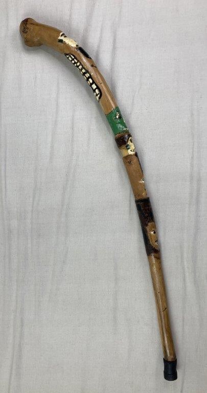 Folk Art Polychromed Carved Cane: Folk art carved wooden cane, signed EH, featuring a curved handle and surface decorated with expressive tribal style faces carved and painted along the shaft. The polychromed detailing highlights the