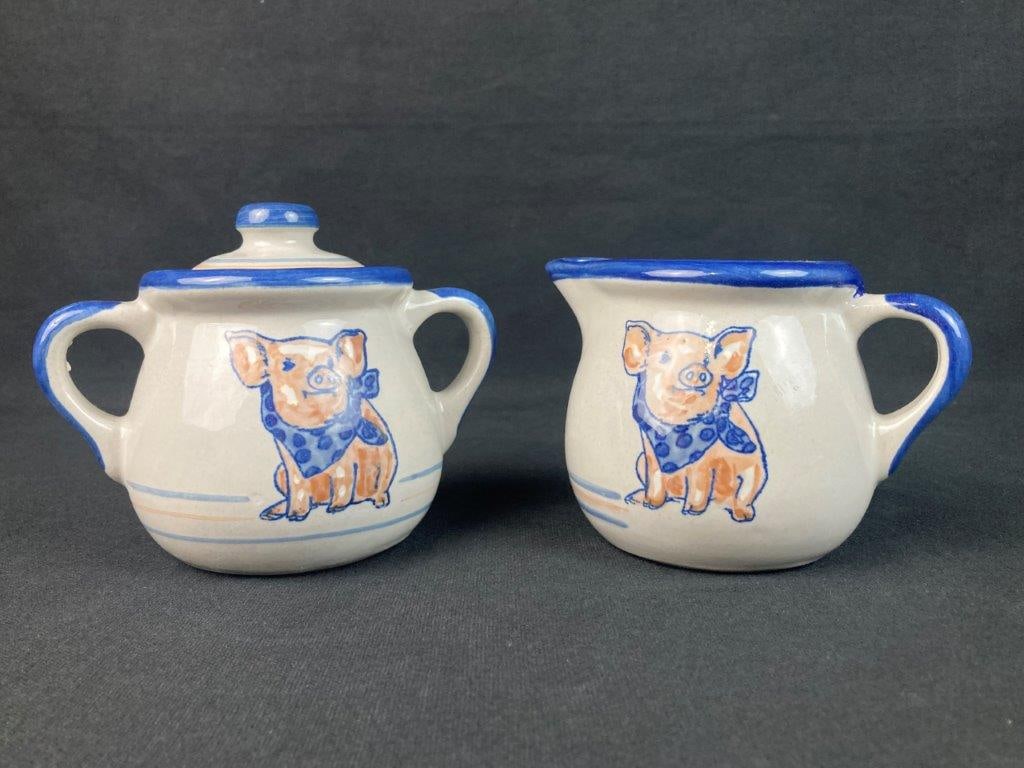 Louisville Stoneware Precious Pig Creamer and Sugar Set: Two-piece set comprising a creamer pitcher and a lidded sugar bowl with dual handles, each decorated with a hand-painted pig wearing a blue polka-dot bandana. The pieces are unified by bold,