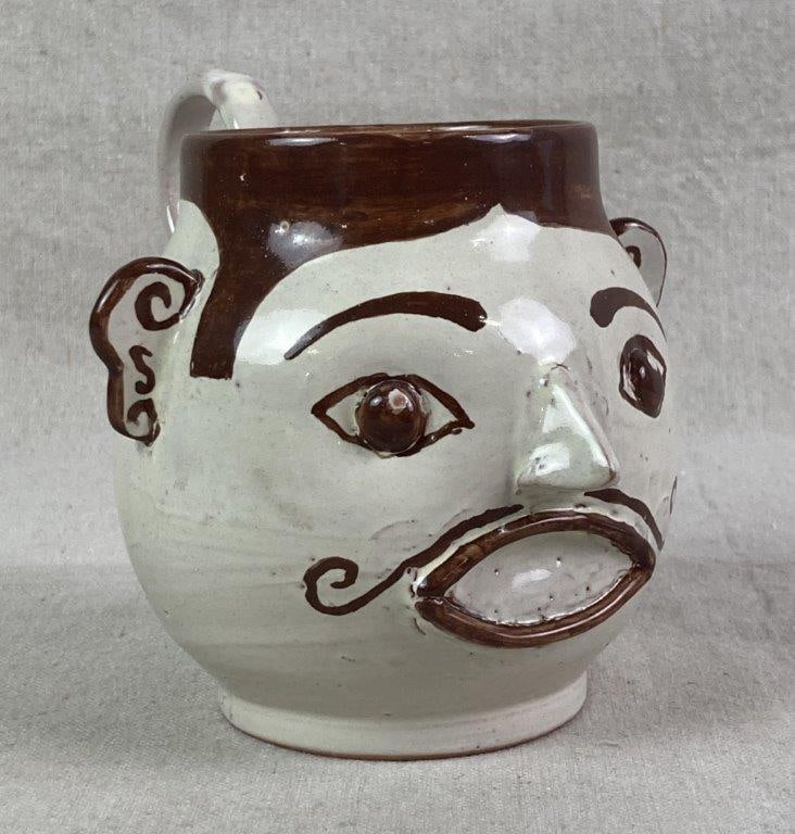 Folk Art Face Mug: White and brown glazed mug featuring an expressive face with a curled mustache and applied ears and nose. Measures approximately 5″ H x 5″ W x 6″ D. Good condition with light wear