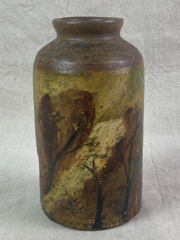 Decorated Stoneware Jar: Cylindrical body decorated with a hand-painted landscape scene depicting a cabin nestled among trees in a naturalistic style with a waterfall. Measures approximately 7 3/4'' H x 4'' W x 4 1/8'' D.