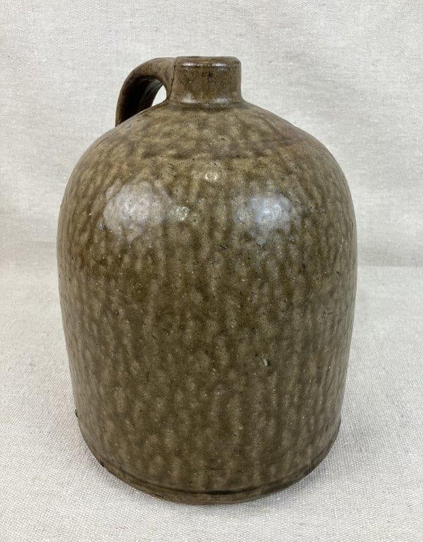 Southern Alkaline Glaze Jug: This stoneware vessel features a curving cylindrical form and an applied strap handle. It is finished in a rich, runny alkaline glaze characteristic of Southern ceramic traditions. The jug measures