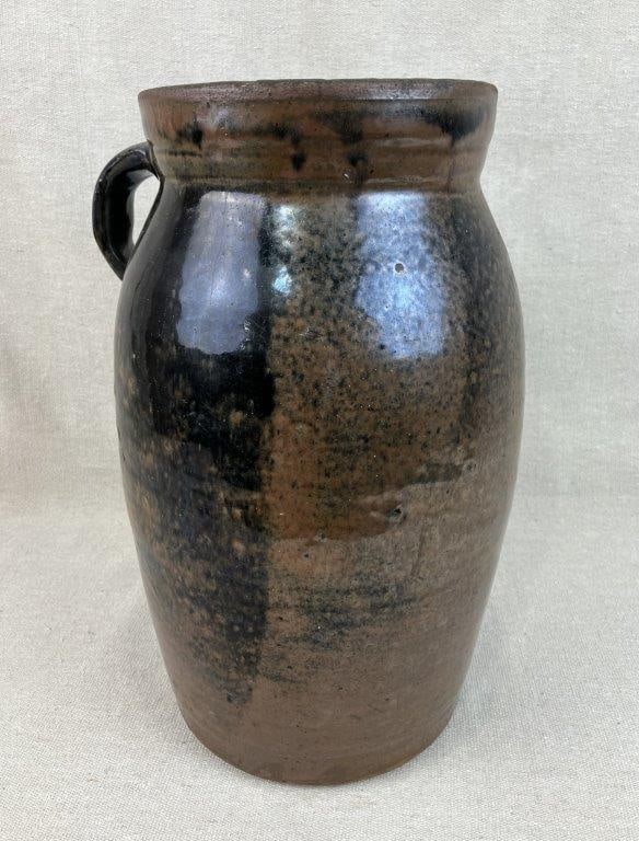 Glazed Stoneware Churn: This vessel features an applied handle and a flared lip finished in a brown glaze with a dark drip effect. It measures approximately 12 7/8" H x 8 1/4" W x 6 1/2" D. The condition includes a visible r