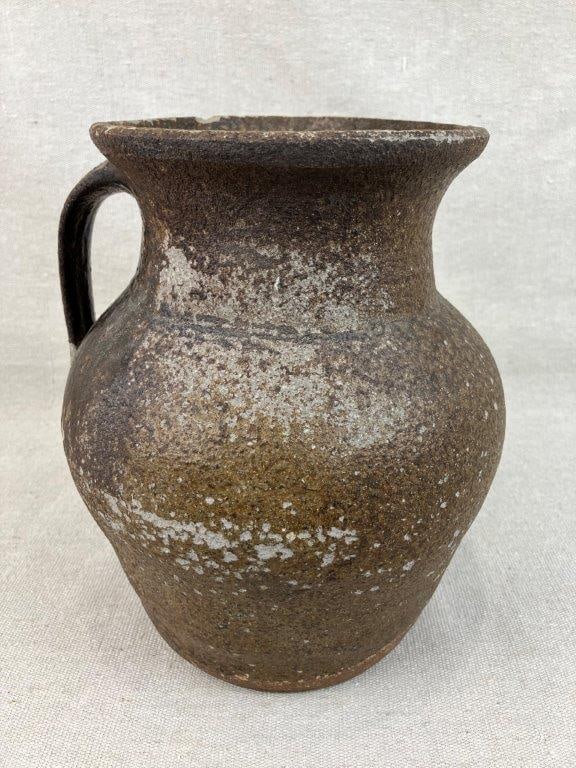 North Carolina Alkaline Glaze Pitcher: This vessel features a traditional curving form with a flared lip and an applied handle, finished in a classic alkaline glaze. Attributed to North Carolina, the piece measures approximately 9 3/8" H