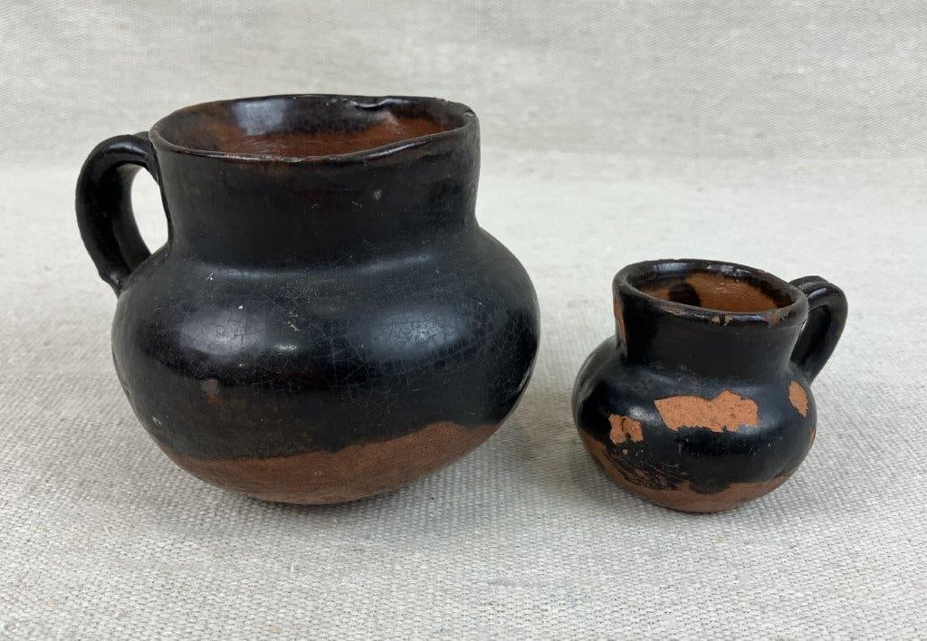 Two Miniature Mexican Black Clay Pottery Pitchers: These delicate vessels, traditionally known as copitas or jarritos, are crafted from a terracotta clay body and finished in a classic black glaze, each featuring a refined form with an applied handle