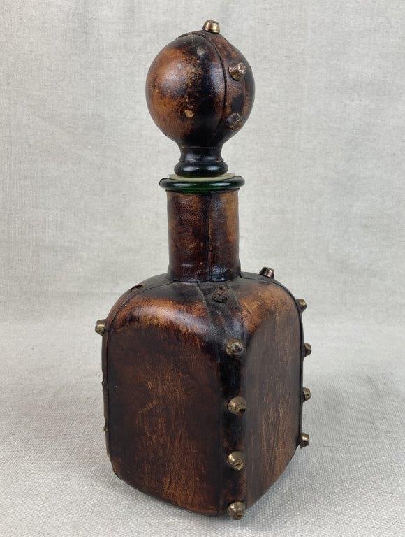Italian Leather Decanter: Leather and glass decanter, likely Italian, featuring brass stud detailing and a globe-form stopper. The surface displays a warm brown tone with natural variation from light to dark, adding visual dep