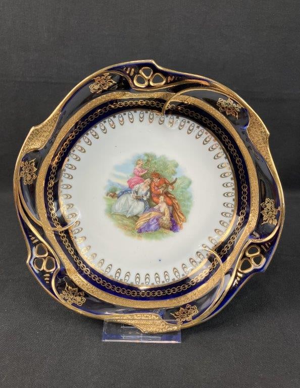 J.L. Menau Gilt Cobalt Painted Porcelain Plate, Germany: Porcelain plate by J.L. Menau, featuring a central Rococo-style pastoral scene with three women and one man in a natural setting, one playing a stringed instrument. The border displays a bold cobalt
