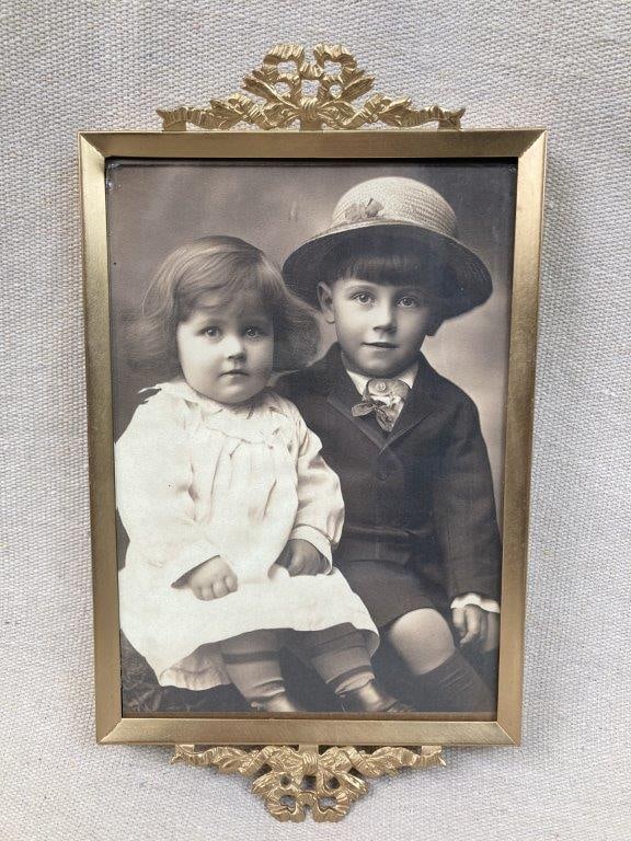 Two Children Framed Photographic Print: Later photographic print depicting two children seated and dressed in early 20th century attire, presented in an ornate, metal gold tone frame. Frame measures approximately 9â€³ H x 5