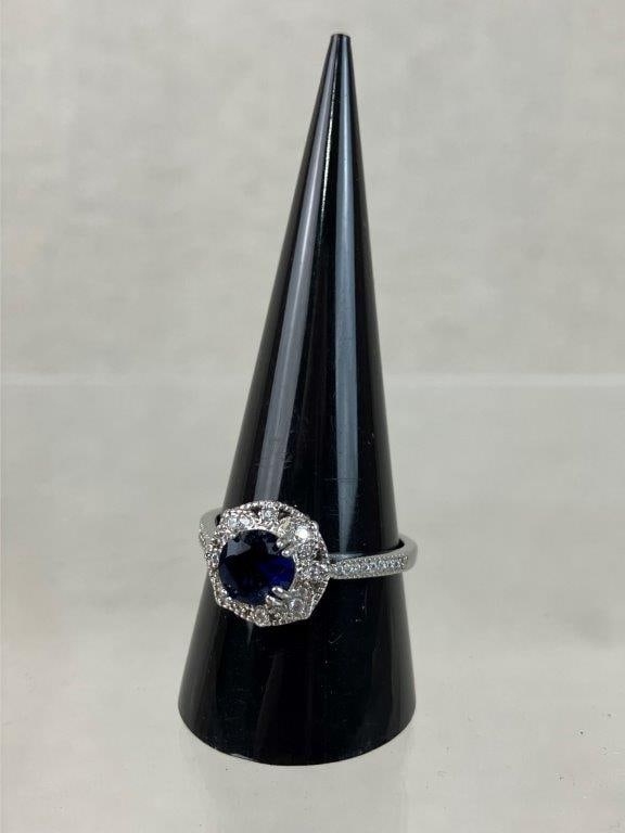 Sterling Silver Sapphire Halo Ring: Sterling silver ring set with a central brilliant cut lab-created sapphire, surrounded by a halo of clear faceted stones within a raised gallery, the band further accented with smaller clear stones al