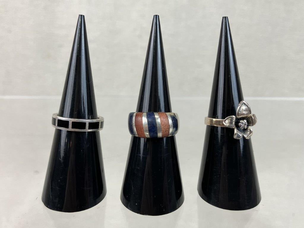 Three Sterling Silver Rings: Group of three sterling silver rings, each marked 925 for silver content, including one with a central lily motif (size approx. 5), one with black onyx inlay (size approx. 5), and one with goldstone