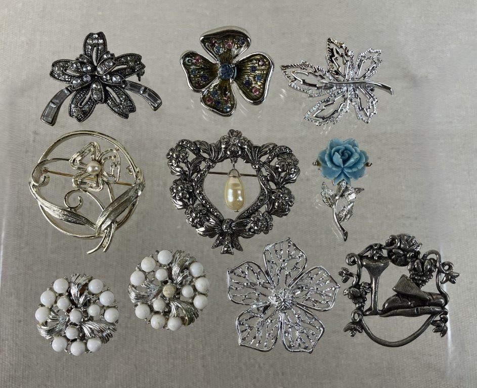 Assortment of Silver-tone Floral Brooches Including Sarah Coventry: This collection of eleven silver-tone brooches and a single pair of clip on earrings features a variety of botanical and unique thematic designs. Highlights include an intricate wreath-style brooch wi