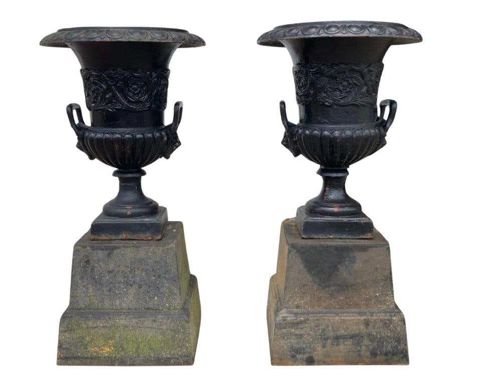 Pair of Ornate Cast Iron Garden Urns: A fine pair of ornate cast iron garden urns featuring flared rims with egg and dart molding, richly decorated with raised floral and foliate motifs, lion mask ring handles, and resting on square