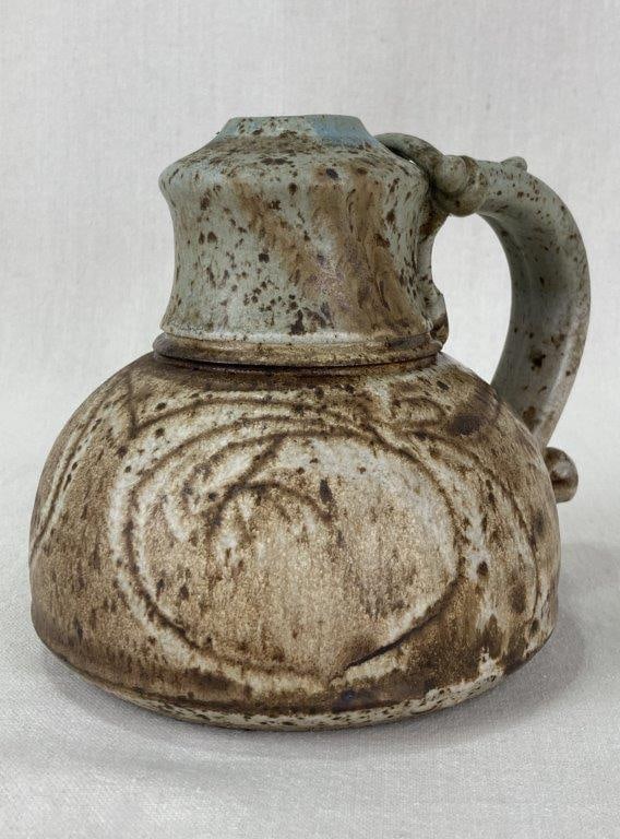 Decorated Studio Pottery Jug: A signed studio pottery pitcher with a bulbous, low-slung body, narrow neck, and small opening. The lower portion showcases an abstract circular pattern finished with an ash-brown glaze, while the