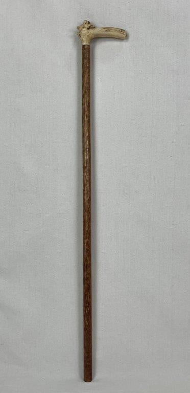 Carved Folk Art Cane: A folk art cane with a straight, polished warm hardwood rod displaying a distinct longitudinal grain pattern and a carved stag horn handle featuring a deer head motif. Measures approximately 35 1/4 in