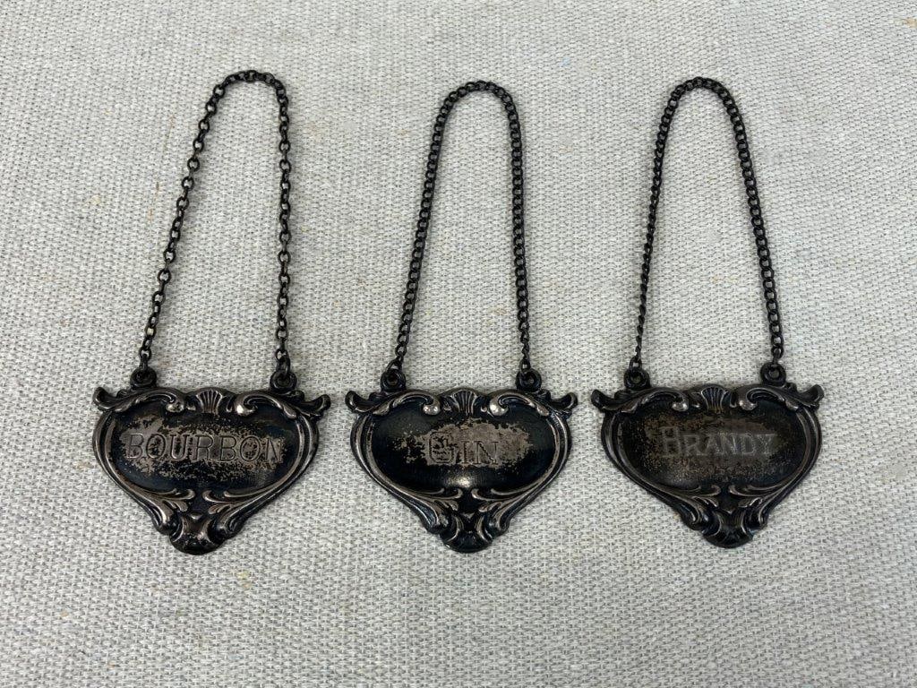 Trio of Sterling Silver Drink Tags: Elegant set of three sterling silver drink tags features individual labels for brandy, bourbon, and gin, each beautifully embellished with sophisticated, classic scrollwork borders that bring a touch 