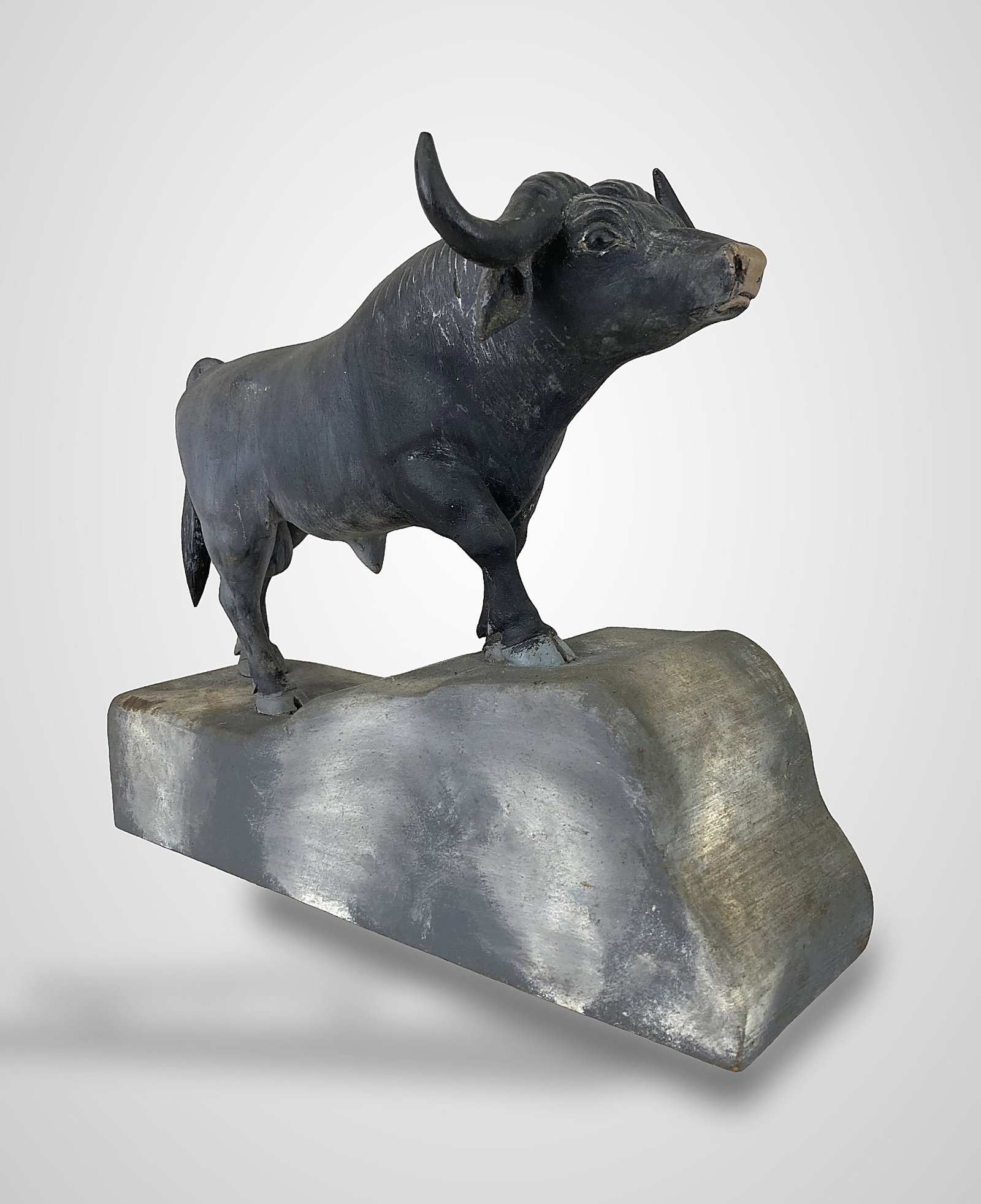 Folk Art Carved Cape Buffalo Statue: This hand-carved wood male Cape Buffalo is mounted to a shaped wood base designed to imitate a rocky outcrop. The buffalo features an alert posture with a raised head, finished in black paint, and is
