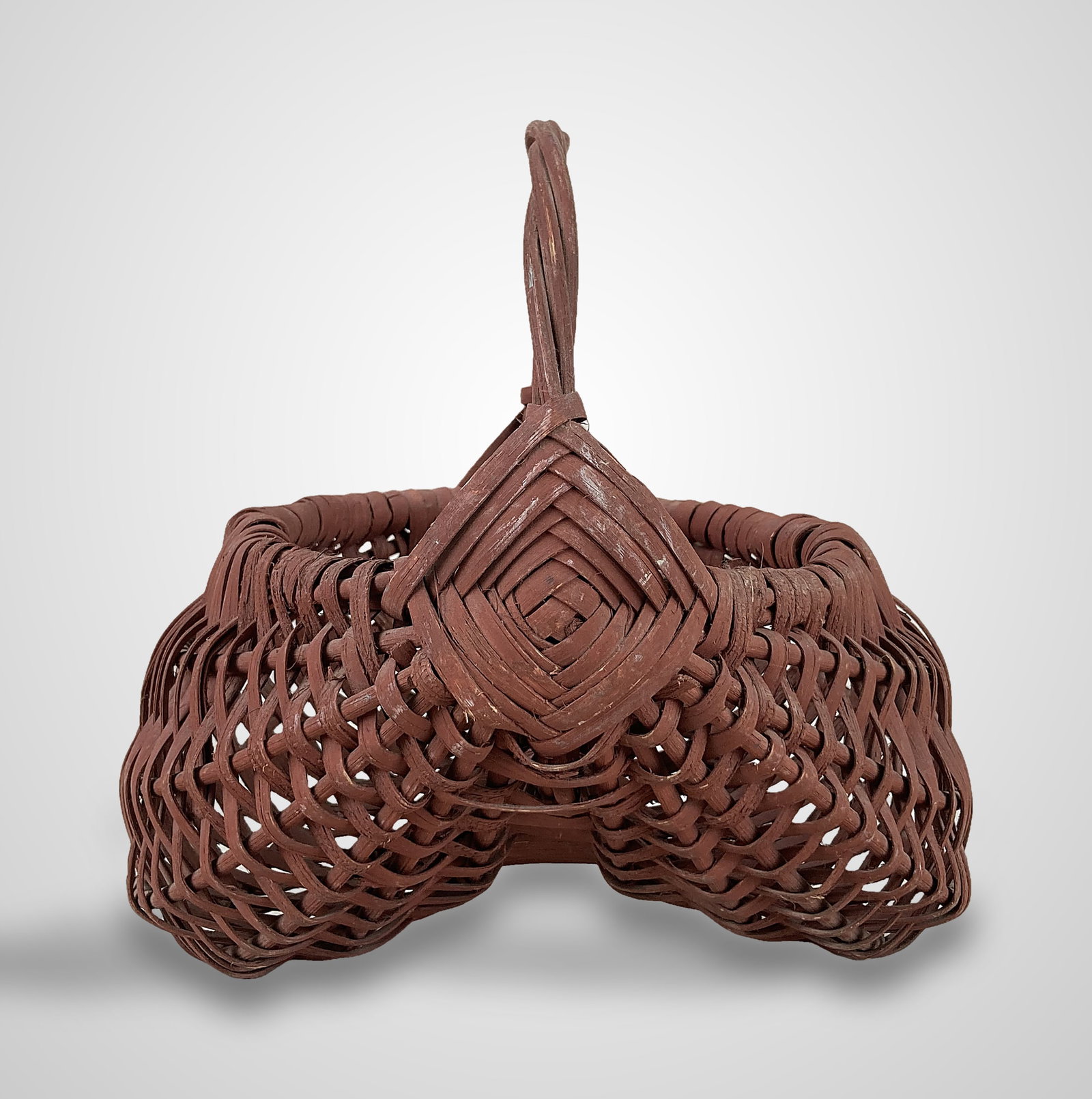 Painted Country Buttocks Basket: This nicely woven split oak buttocks basket is finished in dark-red paint and features a God's eye joint on each side of the handle. It displays a worn patina. It measures approximately 9 1/4 in. H x 