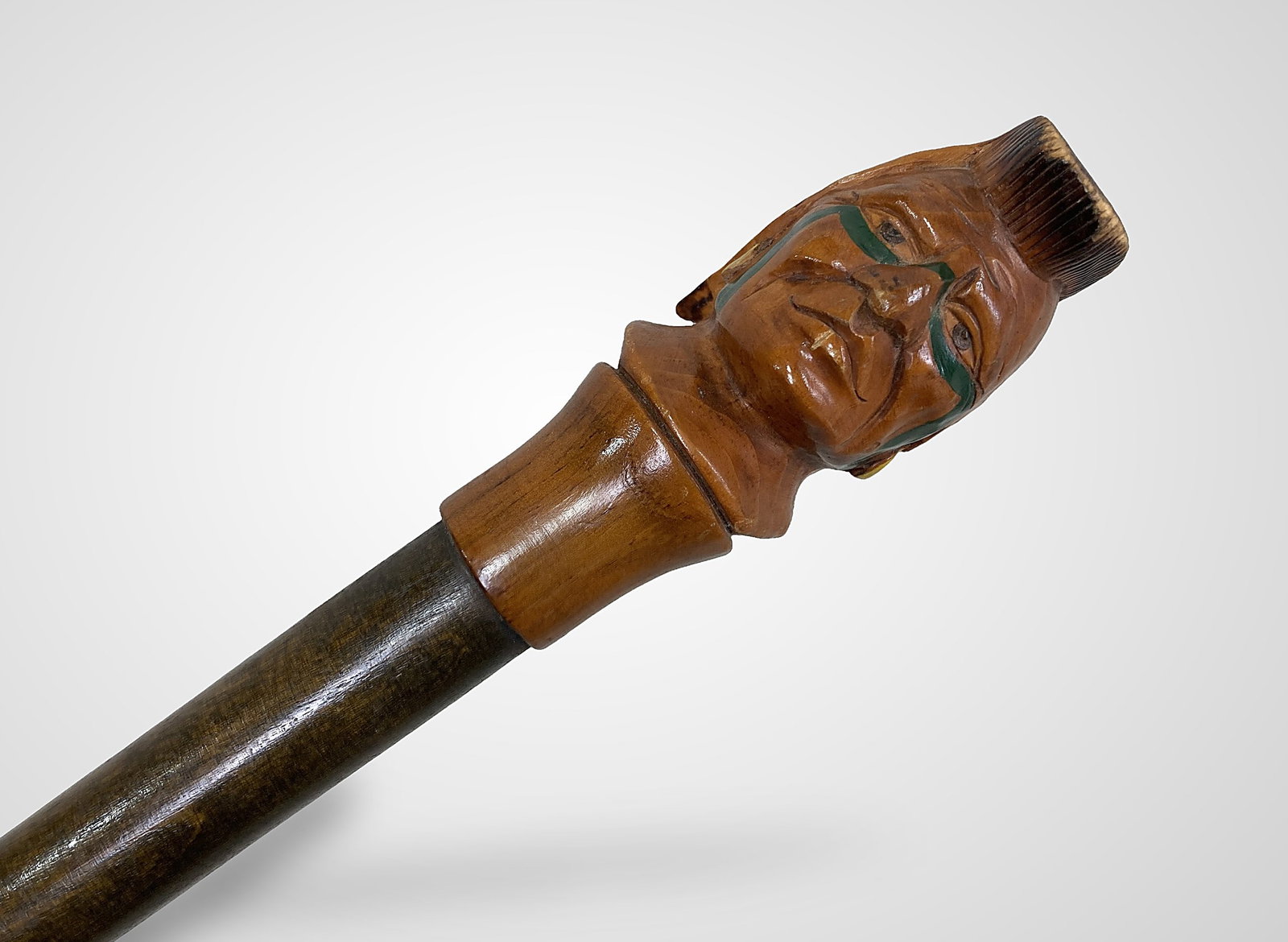 Native American Folk Art Carved Cane: This folk art walking stick features a finely carved handle depicting a male Native American face, complete with green face paint, yellow hoop earrings, and a feathered mohawk. The piece is marked