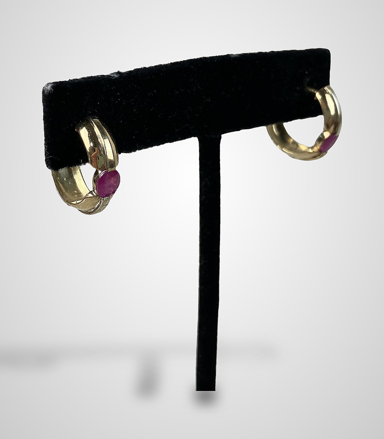 14 KT Gold Ruby Earrings: Huggie hoop earrings are crafted in 14 KT gold and feature a substantial band design. Each earring is set with a central faceted, oval-cut ruby, with a total of 1 CT of ruby weight for the pair. They