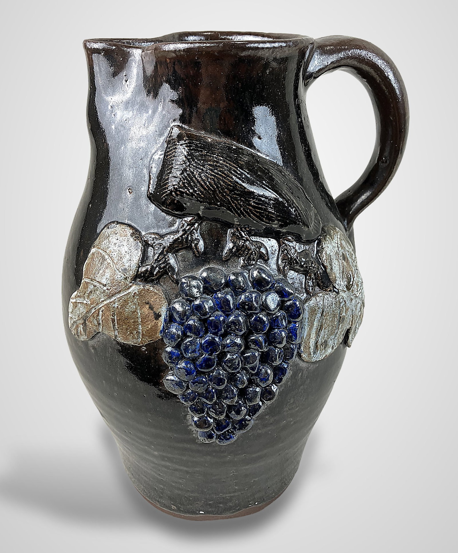 Grace Nell Hewell Southern Stoneware Pitcher: Pitcher features a deep brown glaze, likely an Albany slip or similar, accented by a grape and vine relief motif. The piece is dated 1999 and features an applied handle. Grace Nell Hewell (1933â&
