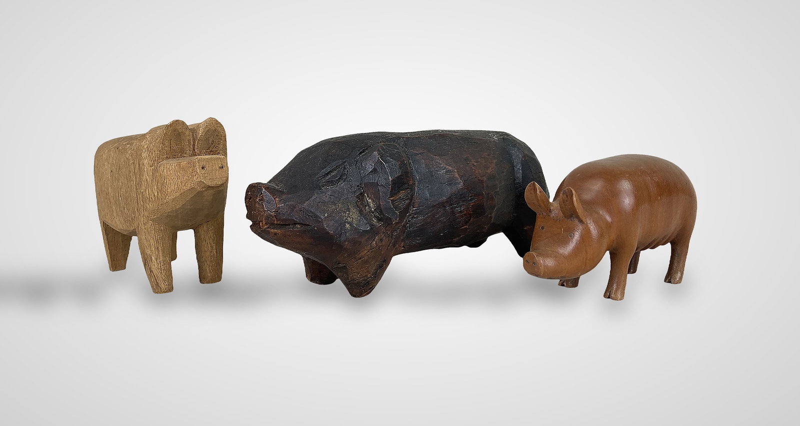 Folk Art Carved Pig Figures: This collection includes three distinct hand-carved wood pig figures. The largest example features crudely carved details with a dark wood finish; a second figure is crafted from polished warm-toned w