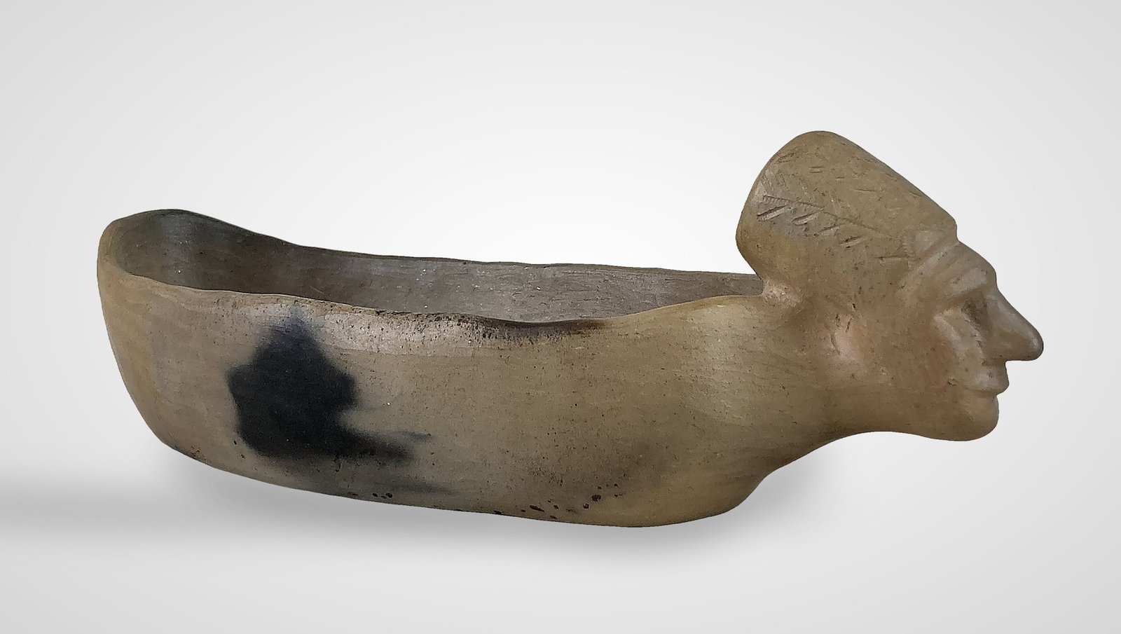 Signed Catawba Indian Canoe Pottery Bowl: A stoneware canoe-shaped bowl featuring a stylized human head on one end with incised foliage details on the headdress. The piece has a tan and brown mottled surface and is signed "Elizabeth Plyler